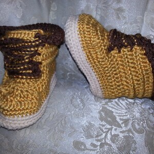 Puede incluir: Un par de botines de bebé tejidos a crochet en marrón, beige y blanco. Los botines tienen un diseño de cordones y un tejido de punto texturizado.