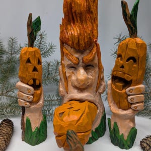 May include: A hand-carved wooden sculpture featuring a central figure with spiky orange hair and prominent eyebrows, holding a carved orange pumpkin in its mouth, flanked by two smaller carved hands each holding a carved orange pumpkin.