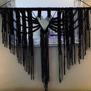 May include: A black macrame wall hanging suspended from a horizontal metal rod with decorative ends. The piece features intricate knotted patterns and long, loose fringe sections that form a V-shaped silhouette, creating a layered texture.