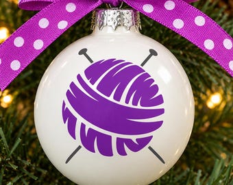 May include: A white spherical ornament featuring a purple graphic of a yarn ball with two crossed knitting needles. The ornament is topped with a silver metal cap and a purple ribbon with white polka dots tied into a decorative bow at the top.