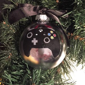 Op de afbeelding: Een zilveren kerstbal met een zwart-wit geverfde videogamecontroller. De controller heeft gekleurde knoppen en een zwart lint.