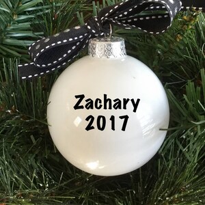 May include: White Christmas ornament with black text that reads "Zachary 2017". The ornament is hanging from a black and white ribbon.