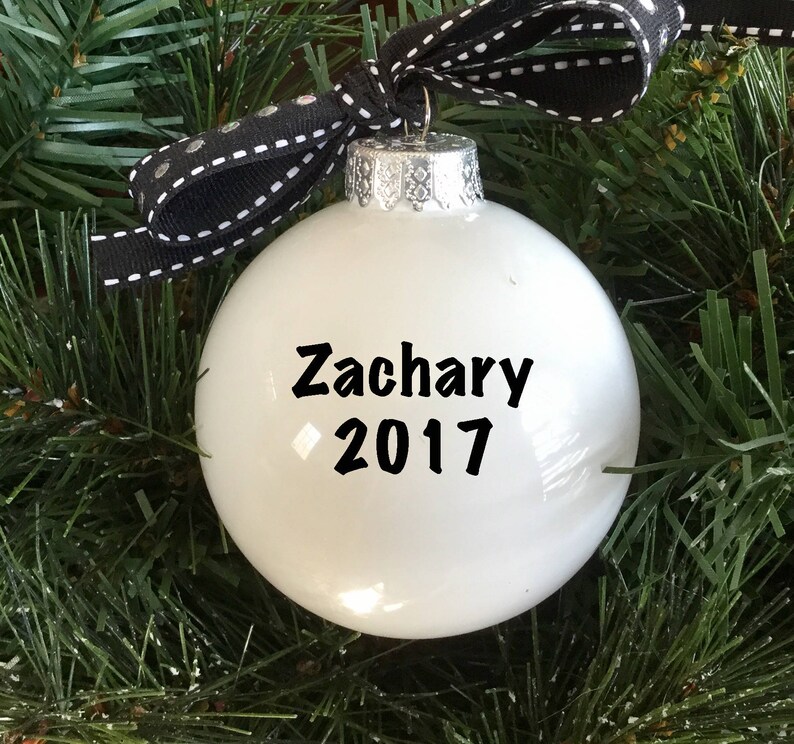 May include: White Christmas ornament with black text that reads "Zachary 2017". The ornament is hanging from a black and white ribbon.