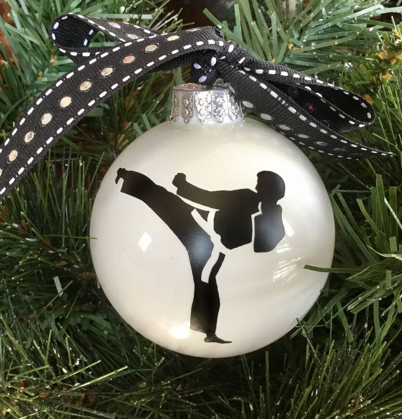 May include: White Christmas ornament with a black silhouette of a person performing a karate kick. The ornament has a silver top and a black and white ribbon.