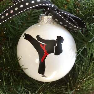 May include: White Christmas ornament with a black silhouette of a person in a karate pose wearing a red belt.