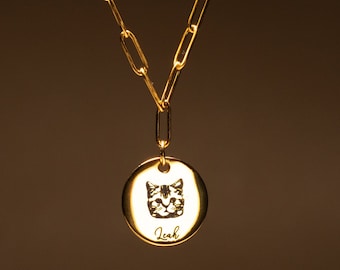 Puede incluir: Collar dorado con una cadena de eslabones grandes y un colgante circular. El colgante presenta una cara de gato grabada y el nombre "Leah" en escritura cursiva. El collar se muestra sobre un fondo marrón cálido.