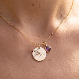 May include: A gold necklace featuring three pendants. One is a disc engraved with "Abigail", and a cross pendant with clear stones. The third pendant is a small, round, purple stone. The necklace is worn around the neck.