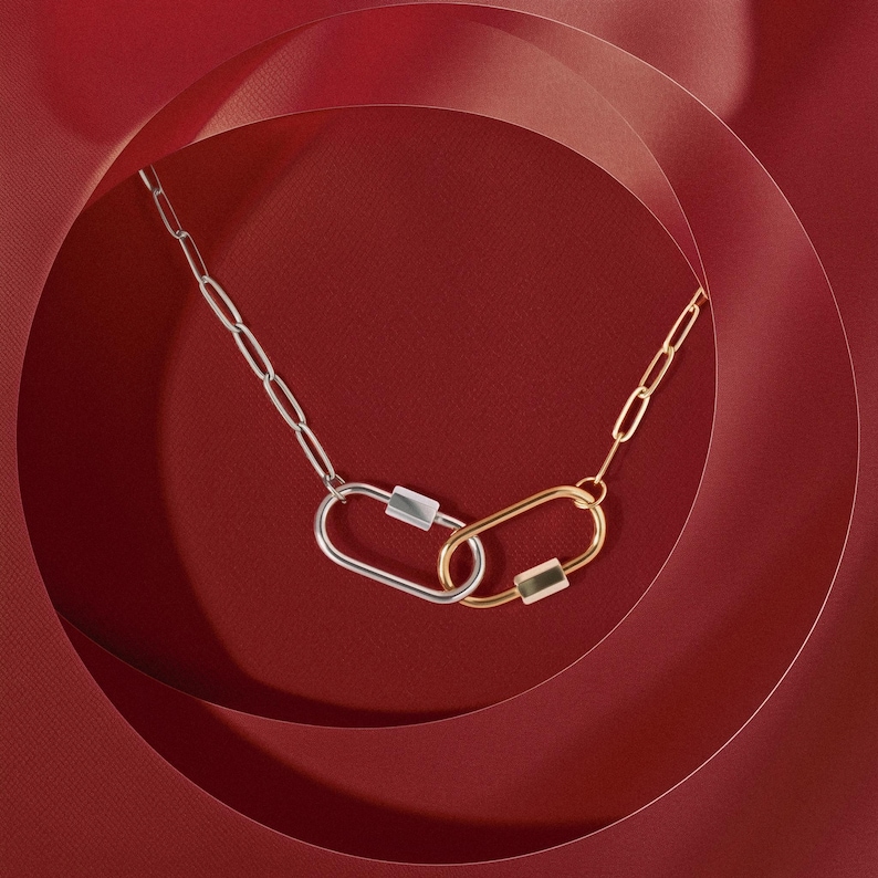 May include: A silver and gold chain necklace with two linked oval-shaped charms. The necklace is photographed on a red background.