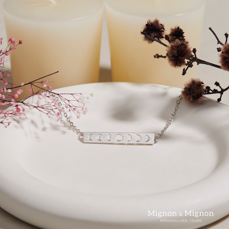 Pode incluir: Um colar de prata com um pingente de barra que apresenta uma s&eacute;rie de recortes em forma de lua crescente. O colar &eacute; exibido em um prato branco com um ramo de flores cor-de-rosa.