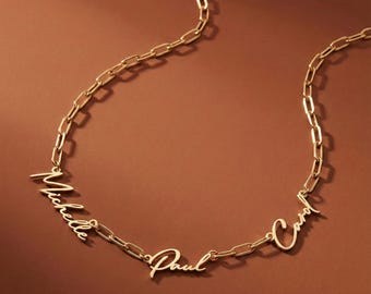 May include: A gold-colored necklace with the names "Michelle", "Paul", and "Carol" in a cursive font. The names are connected by a chain link design. The necklace is displayed on a brown background.