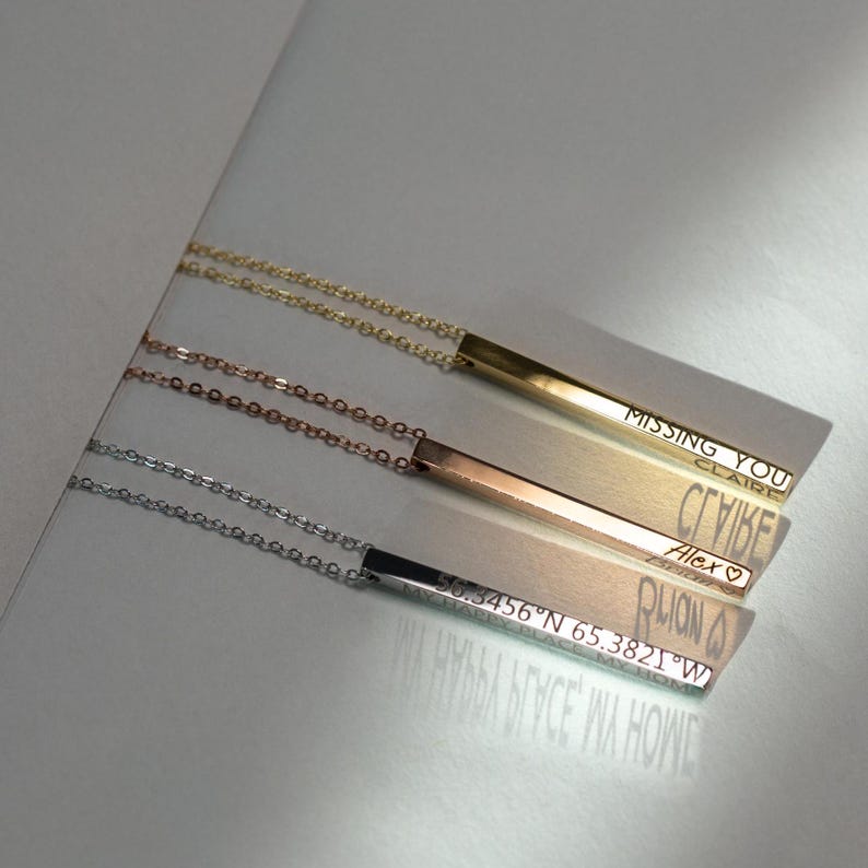 May include: Three gold bar necklaces with different engravings. The top necklace has the words "Missing You Claire" engraved on the bar. The middle necklace has the word "СГУБЕ" engraved on the bar. The bottom necklace has the words "Alex", "PLIAN", and "56.3456&deg;N 65.3821&deg;W" engraved on the bar.