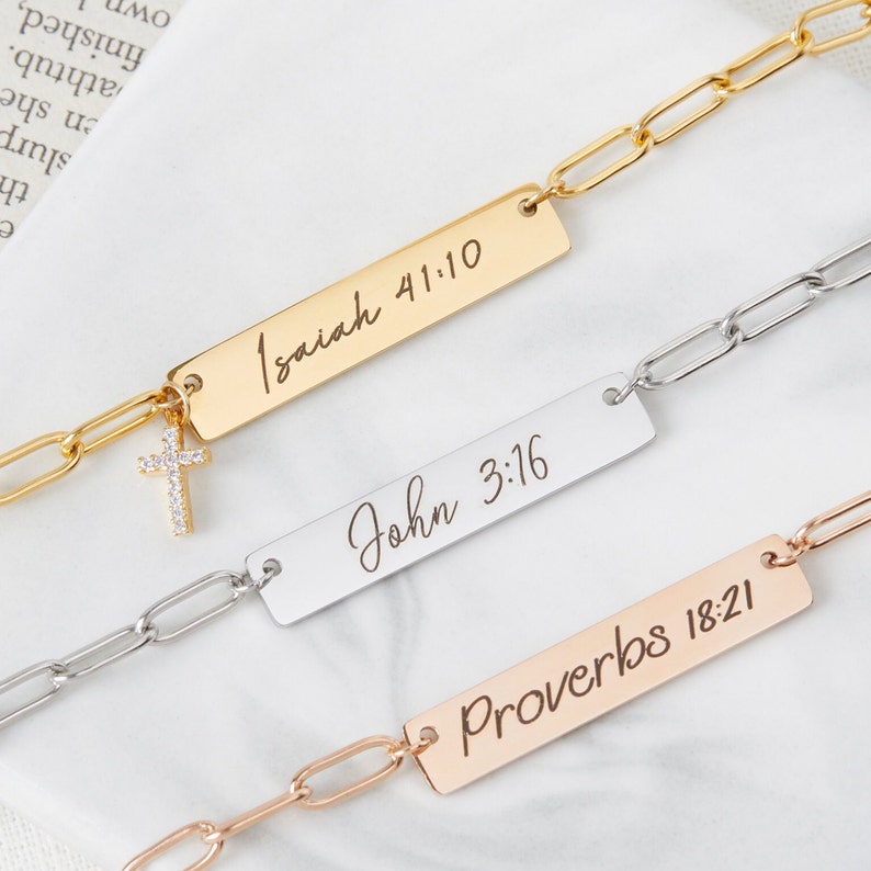 May include: Three chain bracelets in gold, silver and rose gold, each with a rectangular charm. The charms are engraved with the bible verses "Isaiah 41:10", "John 3:16", and "Proverbs 18:21". The gold bracelet features a small cross charm with clear rhinestones.
