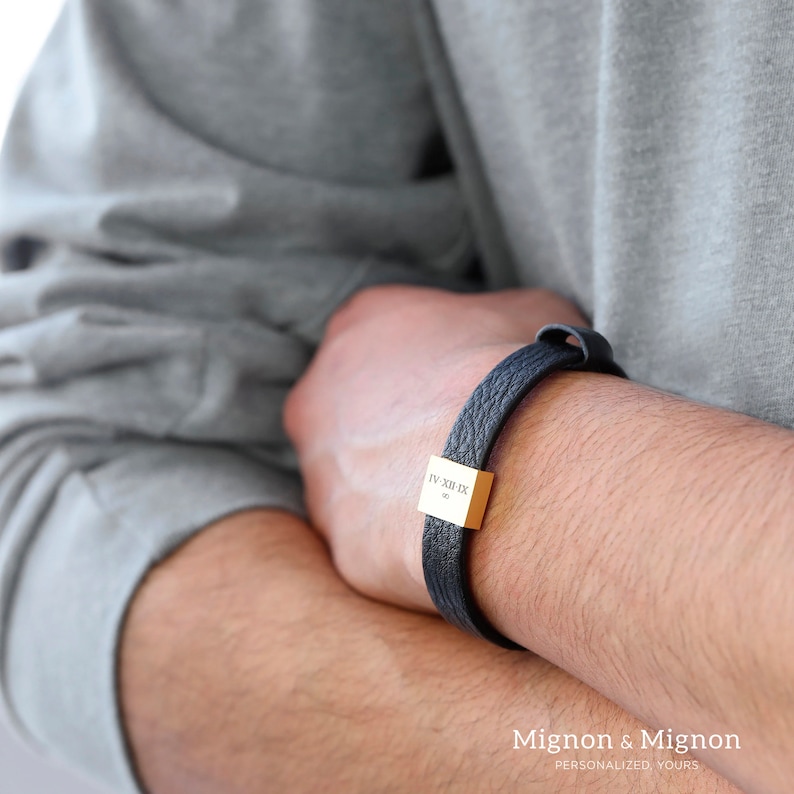 A man in shirt wearing the black leather bracelet that has gold square part with personal engraving on it.