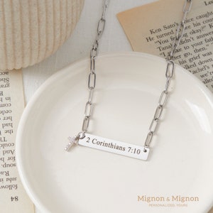 May include: A silver chain necklace with a small cross charm and a rectangular pendant engraved with the text "2 Corinthians 7:10".