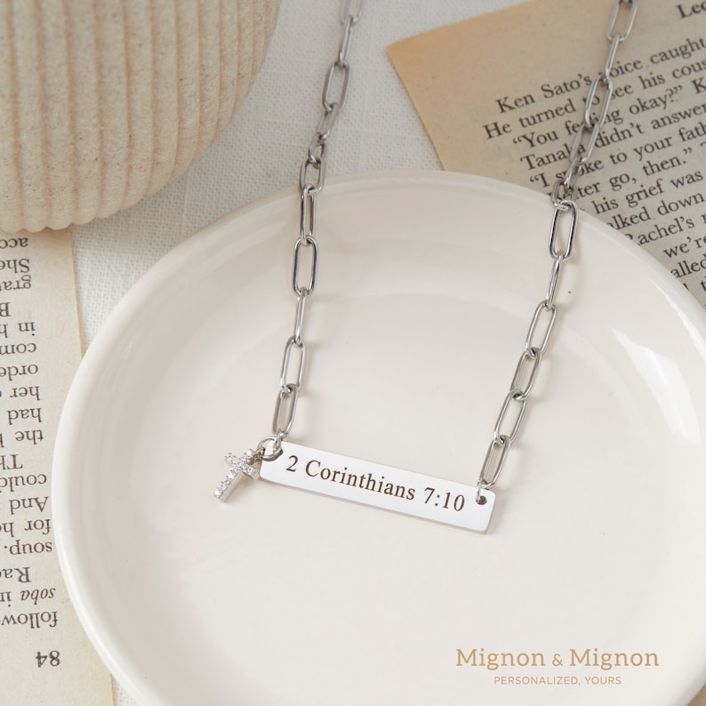 May include: A silver chain necklace with a small cross charm and a rectangular pendant engraved with the text "2 Corinthians 7:10".