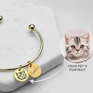 Pode incluir: Pulseira de ouro com dois pingentes. Um pingente tem uma foto em preto e branco de um gato e o outro pingente tem o nome "Simba" gravado.