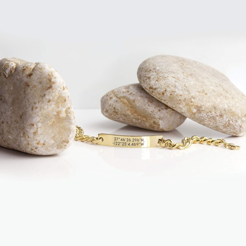 Pode incluir: Pulseira de corrente dourada com uma etiqueta retangular gravada com as coordenadas "37&deg;46'26.296"N -122&deg;25'4.469"W".