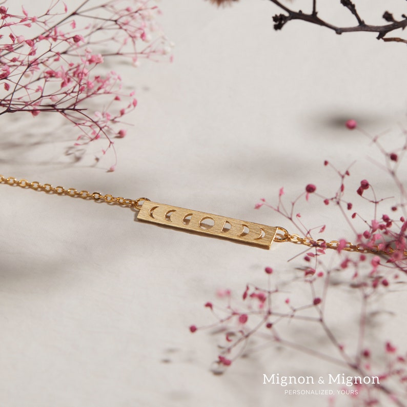 Pode incluir: Um colar de ouro com um pingente de barra que apresenta um design recortado da lua em diferentes fases. O colar est&aacute; sobre um fundo branco com flores cor-de-rosa.