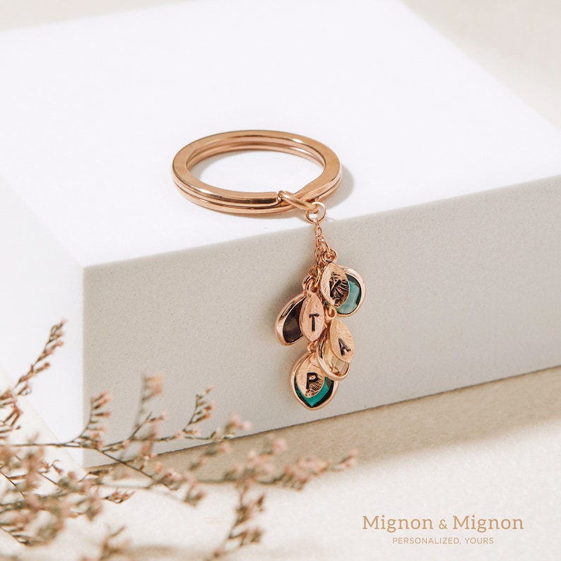 May include: Rose gold keychain with a circular ring and a charm with multiple small teardrop-shaped charms with letters and green and blue stones. The charms spell out "T, A, B, R".