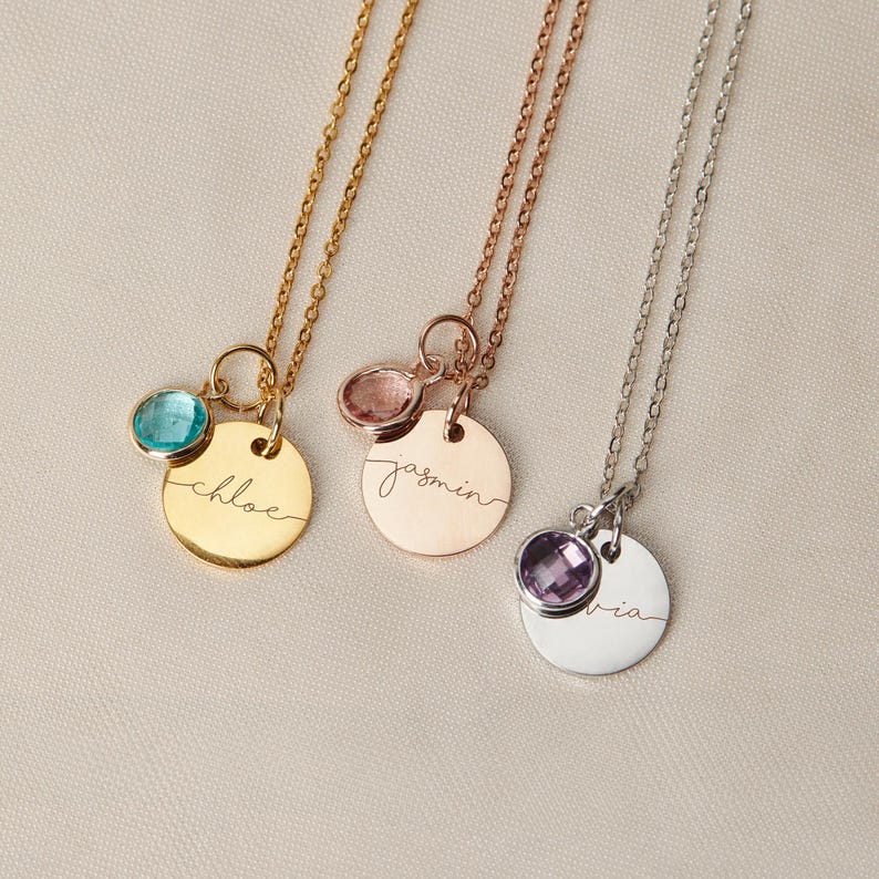 May include: Three gold, rose gold, and silver necklaces with personalized name tags and gemstone charms. The gold necklace has a turquoise charm and the name "Chloe" engraved on the tag. The rose gold necklace has a pink charm and the name "Jasmin" engraved on the tag. The silver necklace has a purple charm and the name "Via" engraved on the tag.