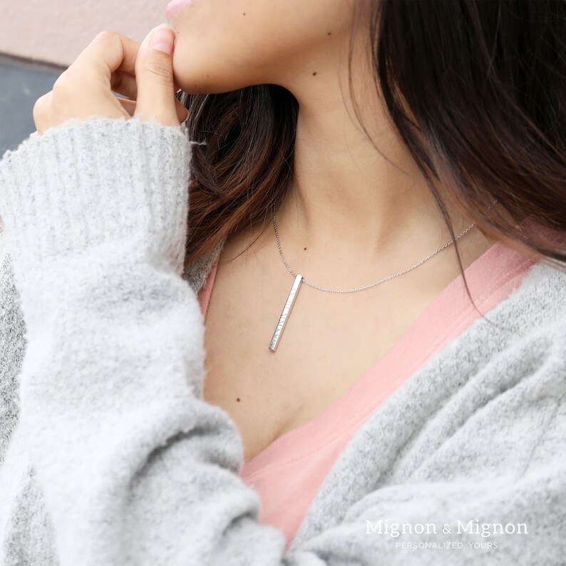 May include: A silver bar necklace with a delicate chain. The necklace is worn by a person with long brown hair and a pink shirt. The text "Mignon & Mignon Personalized Yours" is visible at the bottom of the image.