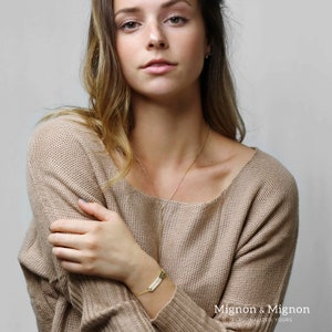 May include: A gold bar bracelet with the words "Mignon & Mignon" engraved on it. The bracelet is on a person's wrist.
