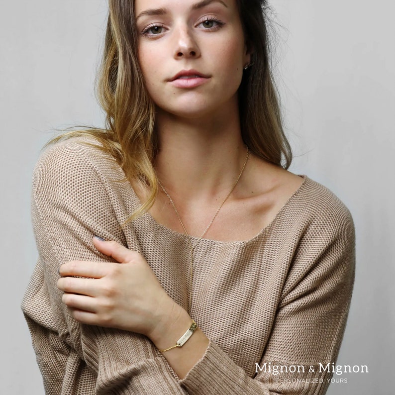 May include: A gold bar bracelet with the words "Mignon & Mignon" engraved on it. The bracelet is on a person's wrist.