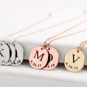 Könnte beinhalten: Drei Halsketten aus Silber, Roségold und Gold mit runden Anhängern. Jeder Anhänger trägt einen gravierten Buchstaben, K, M und V, mit einem Datum darunter. Die Halsketten liegen auf einer weißen Oberfläche.