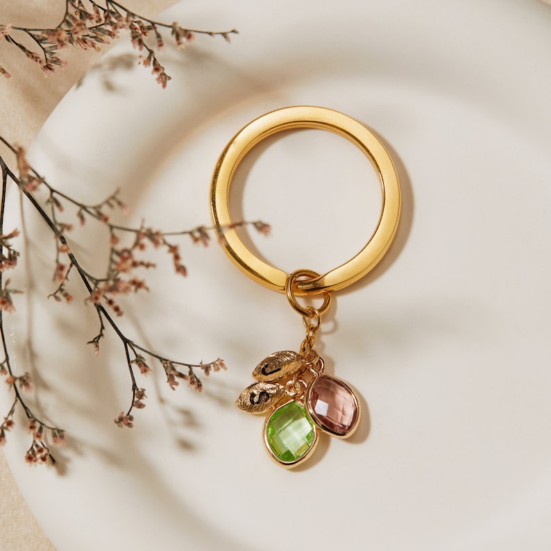 May include: Gold key chain with a round ring and three charms. The charms are a small leaf with a letter on it, a light green gemstone, and a light purple gemstone.