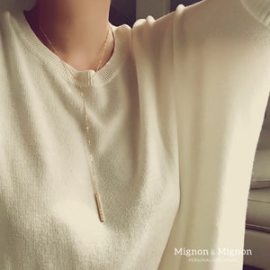 May include: A long, thin gold necklace with a small, rectangular pendant. The necklace is worn by a person wearing a white sweater. The text "Mignon & Mignon" is visible at the bottom of the image.