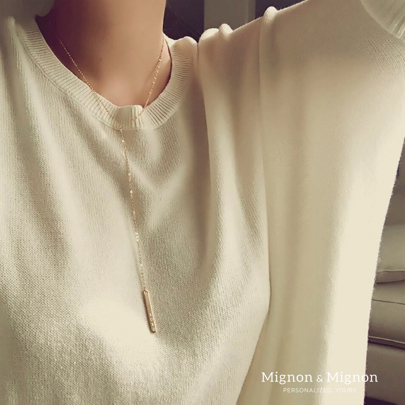May include: A long, thin gold necklace with a small, rectangular pendant. The necklace is worn by a person wearing a white sweater. The text "Mignon & Mignon" is visible at the bottom of the image.