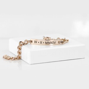 Pode incluir: Um bracelete dourado com um design de corrente de elos e uma etiqueta retangular gravada com os numerais romanos "IV. X. MMXIV".
