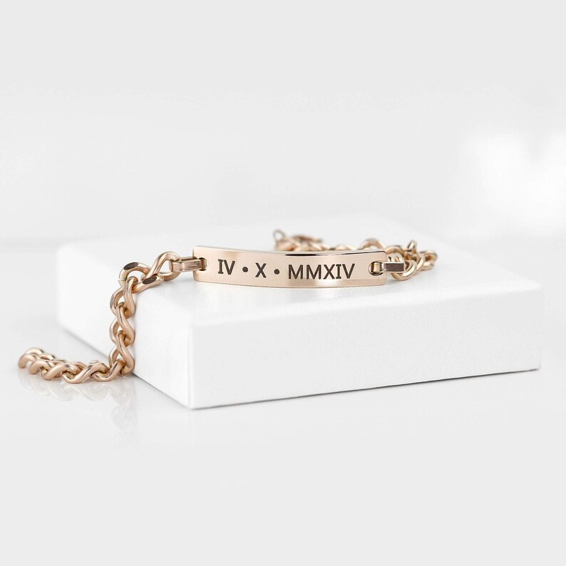 Pode incluir: Um bracelete dourado com um design de corrente de elos e uma etiqueta retangular gravada com os numerais romanos "IV. X. MMXIV".