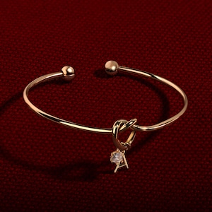 Puede incluir: Una pulsera dorada con un diseño de nudo y un pequeño colgante de piedra preciosa transparente. La pulsera tiene extremos redondeados en forma de bola y se muestra sobre una superficie roja texturizada.