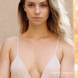 May include: A gold chain necklace with a long, thin pendant hangs from the neck of a person wearing a beige tank top. The pendant is a thin, rectangular bar. The text "Mignon & Mignon" is visible at the bottom of the image.