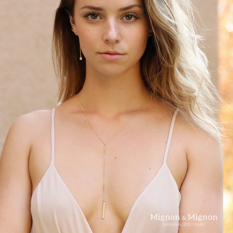 May include: A gold chain necklace with a long, thin pendant hangs from the neck of a person wearing a beige tank top. The pendant is a thin, rectangular bar. The text "Mignon & Mignon" is visible at the bottom of the image.
