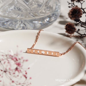Pode incluir: Um colar de ouro rosa com um pingente de barra que apresenta um design recortado de seis fases lunares. O colar est&aacute; sobre um prato branco com um padr&atilde;o floral.