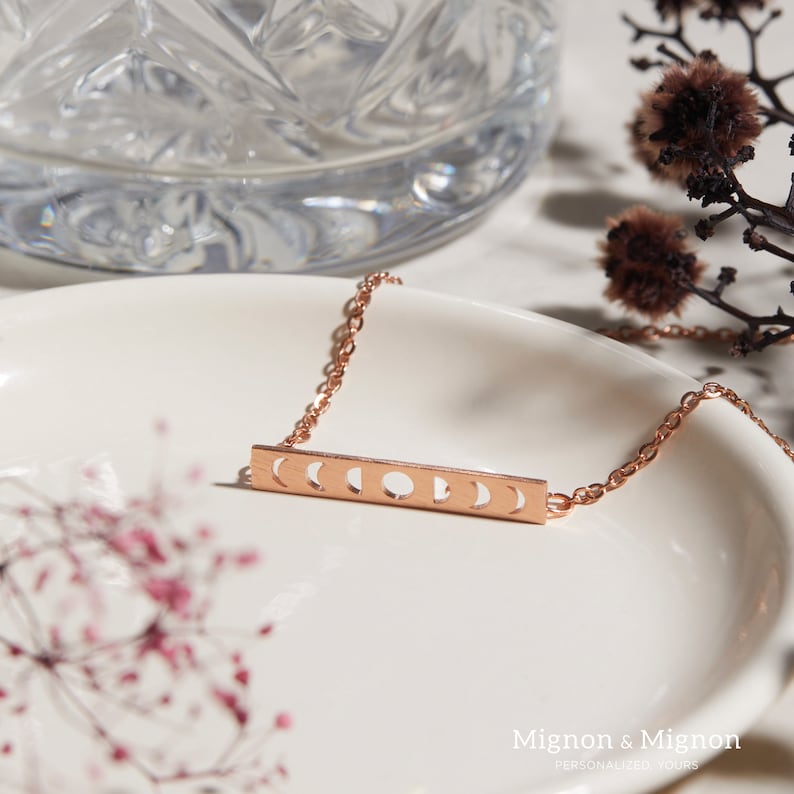 Pode incluir: Um colar de ouro rosa com um pingente de barra que apresenta um design recortado de seis fases lunares. O colar est&aacute; sobre um prato branco com um padr&atilde;o floral.