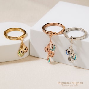 May include: Three gold keychains with charms. Each charm has a different colored gemstone and a letter engraved on it. The keychains are on a white surface. Mignon & Mignon Personalized. Yours.