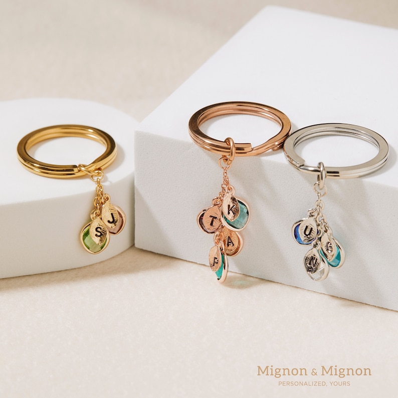 May include: Three gold keychains with charms. Each charm has a different colored gemstone and a letter engraved on it. The keychains are on a white surface. Mignon & Mignon Personalized. Yours.