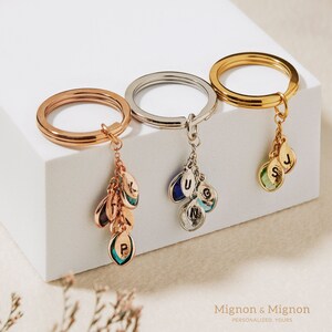 May include: Three gold keychains with charms. Each keychain has a different colored ring: rose gold, silver, and gold. The charms are small teardrop-shaped pendants with letters engraved on them. The charms are attached to the keychains with small chains.