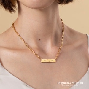 May include: A gold chain necklace with a rectangular pendant that says "Nahum 1:7" in cursive script.