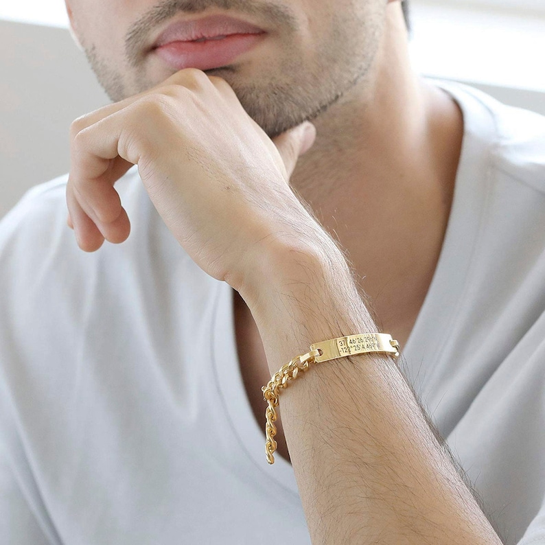 Pode incluir: Um bracelete de corrente de ouro com uma etiqueta retangular gravada com uma s&eacute;rie de n&uacute;meros. O bracelete est&aacute; no pulso de uma pessoa.