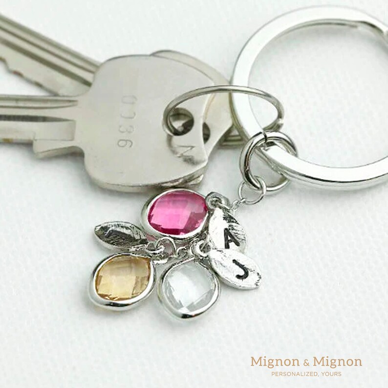 May include: A silver keychain with a charm featuring four small crystals in pink, yellow, and clear, and two silver charms with the letters A and J engraved on them. The keychain is attached to a silver key ring.