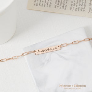May include: A rose gold chain bracelet with a rectangular charm that reads "Proverbs 18:21".