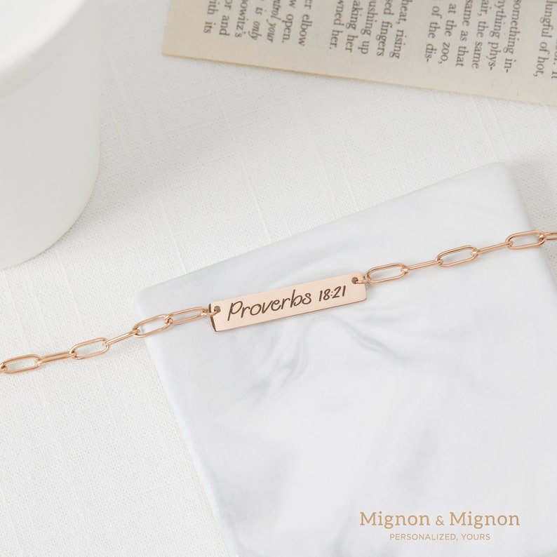 May include: A rose gold chain bracelet with a rectangular charm that reads "Proverbs 18:21".