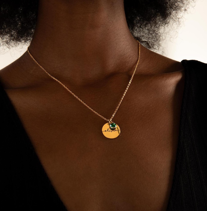May include: A gold necklace with a round pendant that says "alive" in cursive script. A small green gemstone is attached to the pendant.