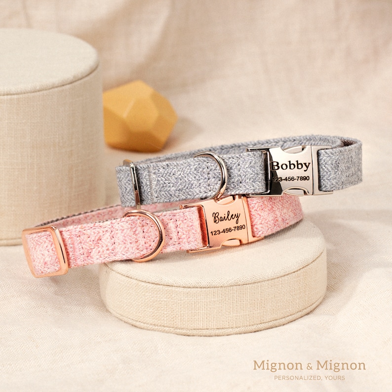 May include: Two personalized dog collars, one pink and one gray, with silver and rose gold buckles. The collars have the names "Bailey" and "Bobby" and phone numbers on the tags. The collars are on a beige fabric surface. Mignon & Mignon Personalized, Yours.