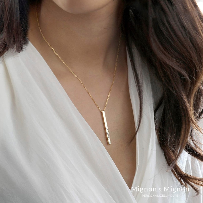 May include: A gold chain necklace with a long, thin, vertical bar pendant. The bar has a textured surface.