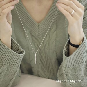 May include: A silver necklace with a long, thin pendant set with small, clear stones. The pendant is a vertical bar shape. The necklace is worn by a person wearing a green sweater.